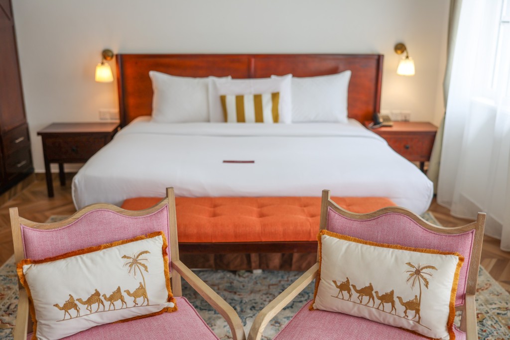 A made bed between two timber bedside tables, with a pair of armchairs at its foot.