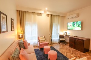 A Deluxe Room with sofa and ottomans, writing desk, wall-mounted television and garden window.