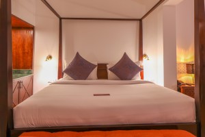 A wide four-poster bed dressed in white linen with dark cushions.