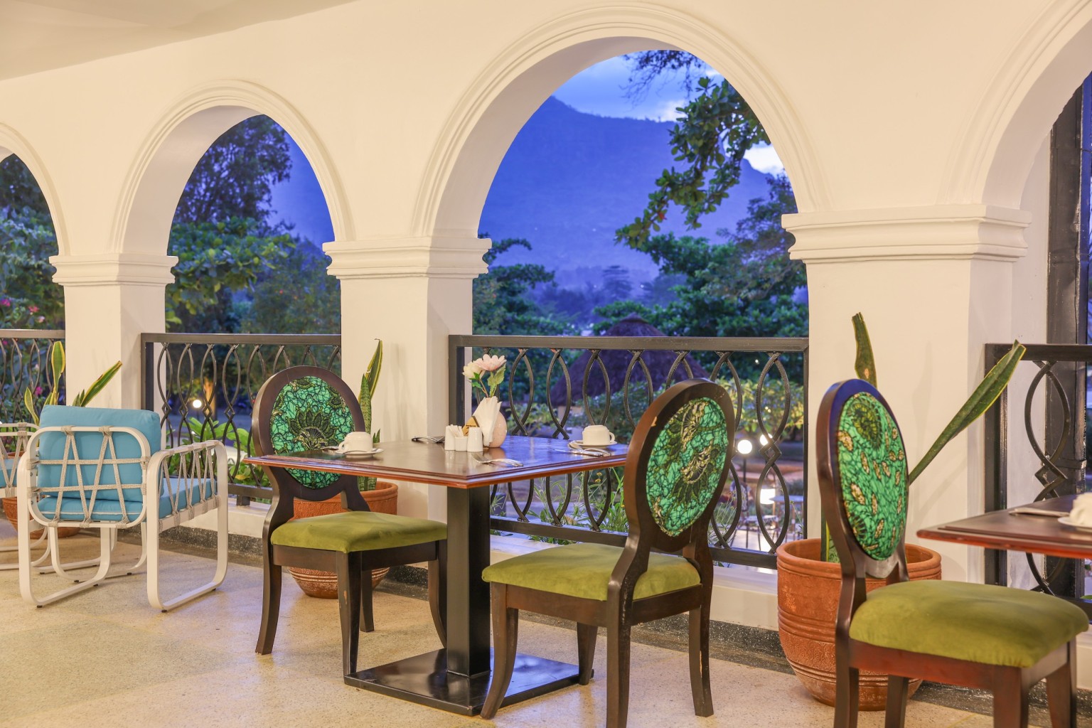Dining tables and cane chairs along the arched veranda, hills visible through the openings.