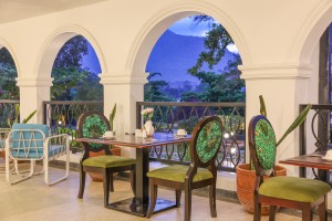 Dining tables and cane chairs along the arched veranda, hills visible through the openings.