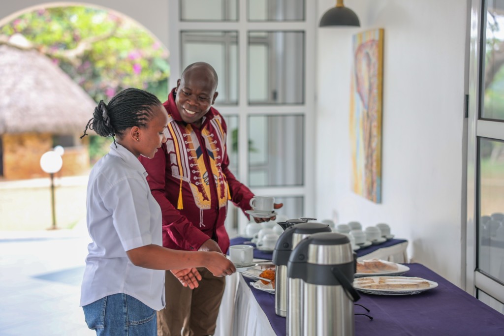 Two delegates at the tea break table, one serving from a covered dish, flasks and cups set out beside them.
