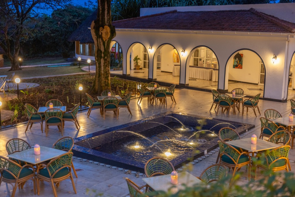 Courtyard tables and cane chairs set around a lit water feature at dusk, the arcades glowing behind.