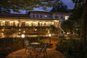 The lit arcaded frontage of the hotel at dusk, seen across the lawns and garden paths.