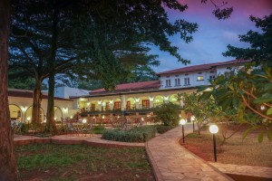 The lit arcaded frontage of the hotel at dusk, seen across the lawns and garden paths.