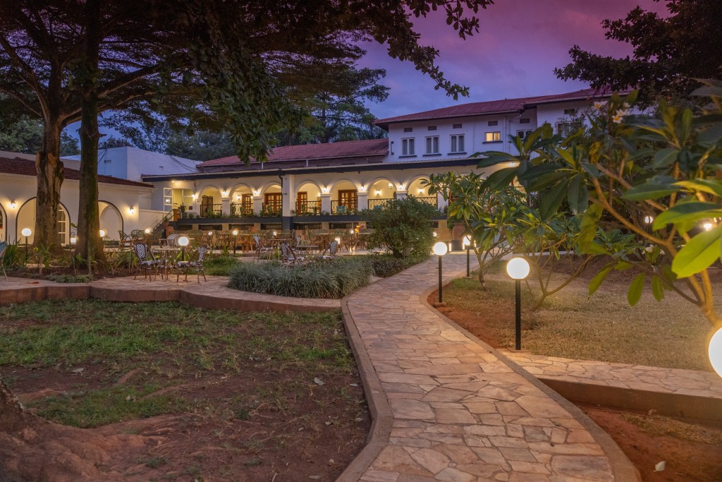 A paved garden path curving between mature trees at dusk, the hotel’s lit arcades along the far side.