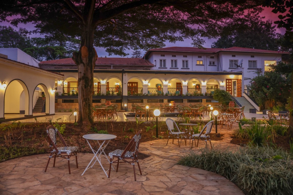 Garden seating on a paved circle under a mature tree, the hotel’s lit arcades along the far side at dusk.