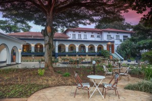 The lit arcaded frontage of the hotel at dusk, seen across the lawns and garden paths.
