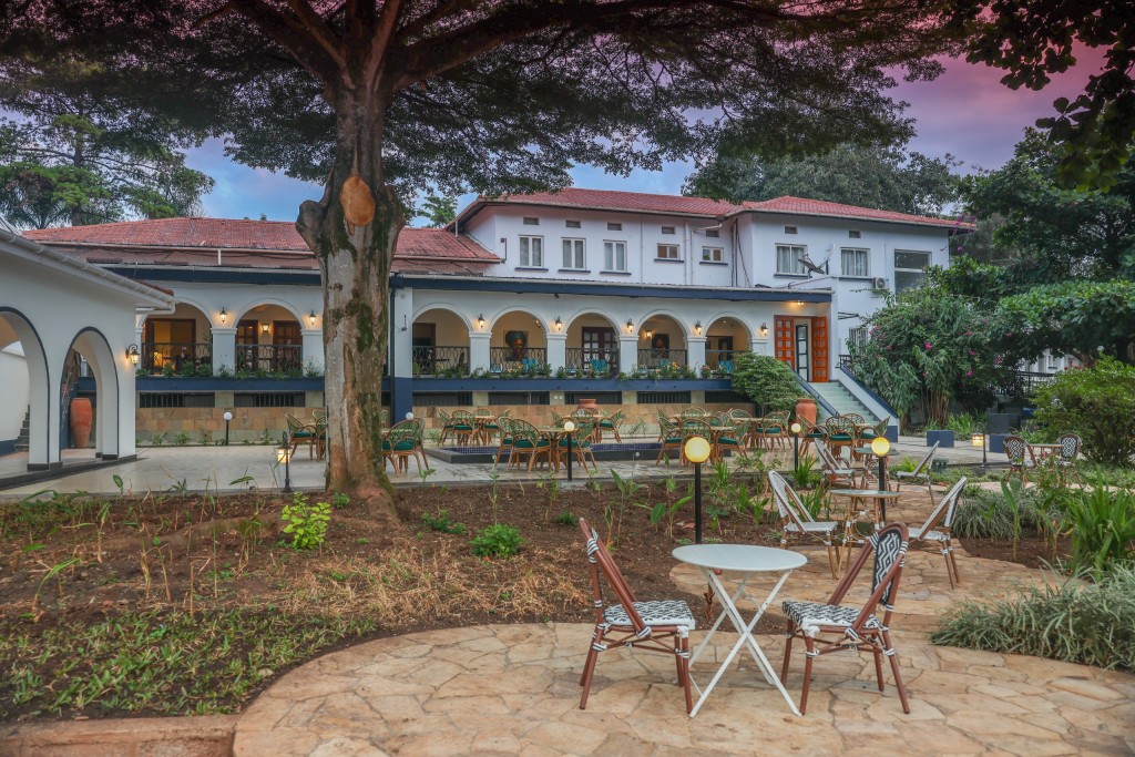 Garden tables and chairs on a paved circle beneath a large tree, with the arcaded frontage behind at dusk.