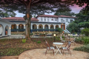 The lit arcaded frontage of the hotel at dusk, seen across the lawns and garden paths.
