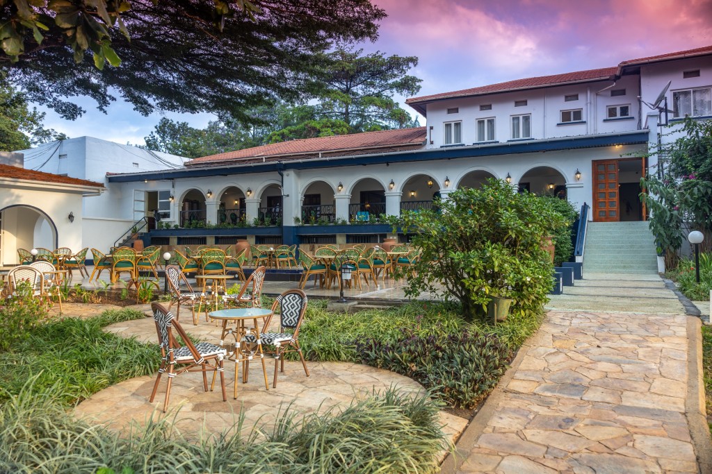 The hotel’s arcaded two-storey wings lit at dusk, seen across the lawn past garden tables and a stone path.