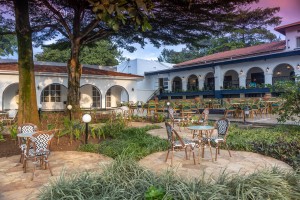 The lit arcaded frontage of the hotel at dusk, seen across the lawns and garden paths.