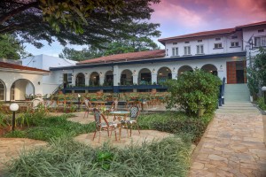 The lit arcaded frontage of the hotel at dusk, seen across the lawns and garden paths.