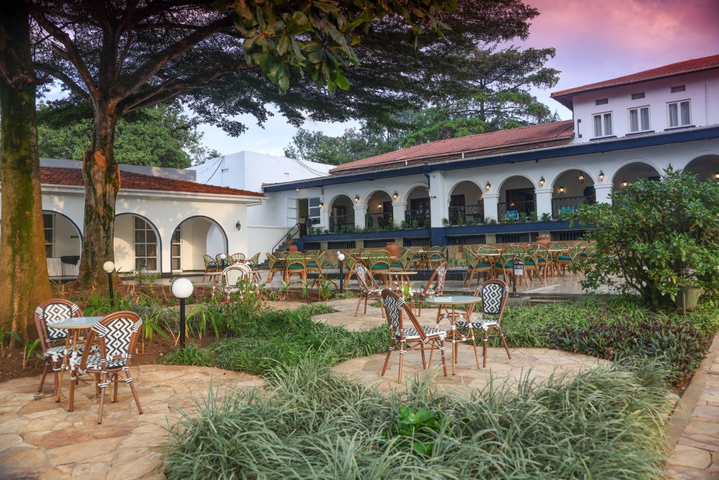 Garden seating under a mature tree with the hotel’s lit arcades and terrace behind it at dusk.