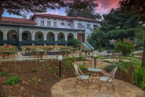 The lit arcaded frontage of the hotel at dusk, seen across the lawns and garden paths.