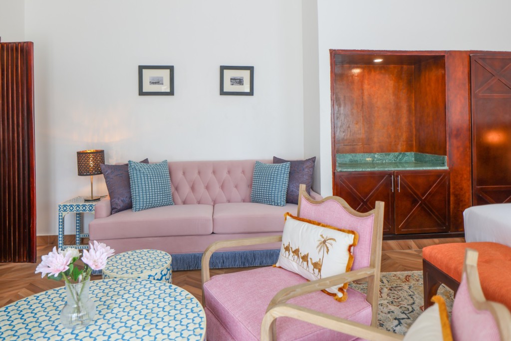 A guest room seating area with a pink sofa, an armchair, a patterned round table and a tall timber wardrobe.