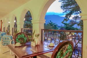 Masaaba Restaurant: tables and cane chairs along the arched veranda, hills and greenery through the openings.