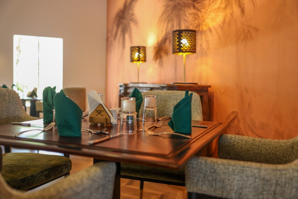 A dining table laid with green napkins under warm wall lamps, the painted palm mural behind it.
