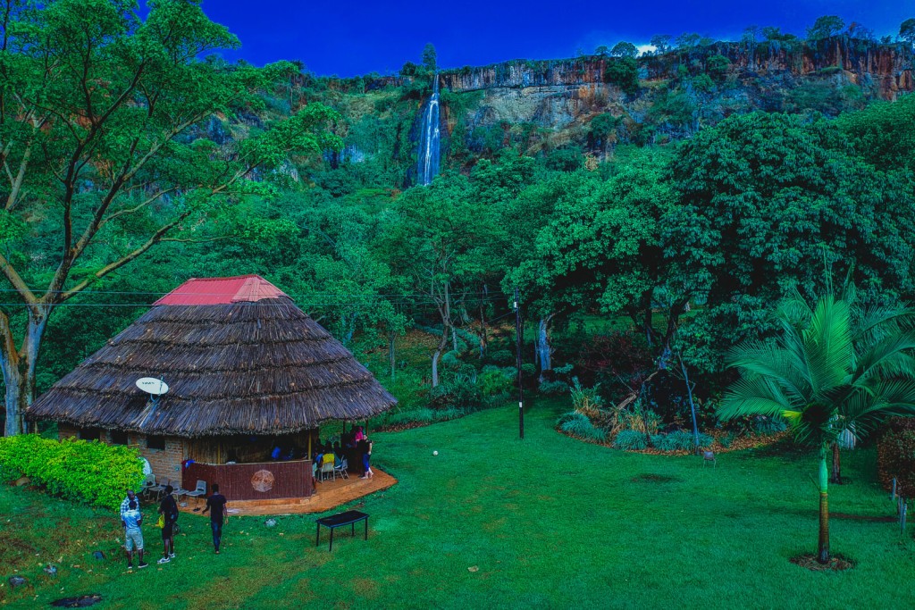 A thatched banda on green lawn with the Sisiyi waterfall dropping down the cliff behind.